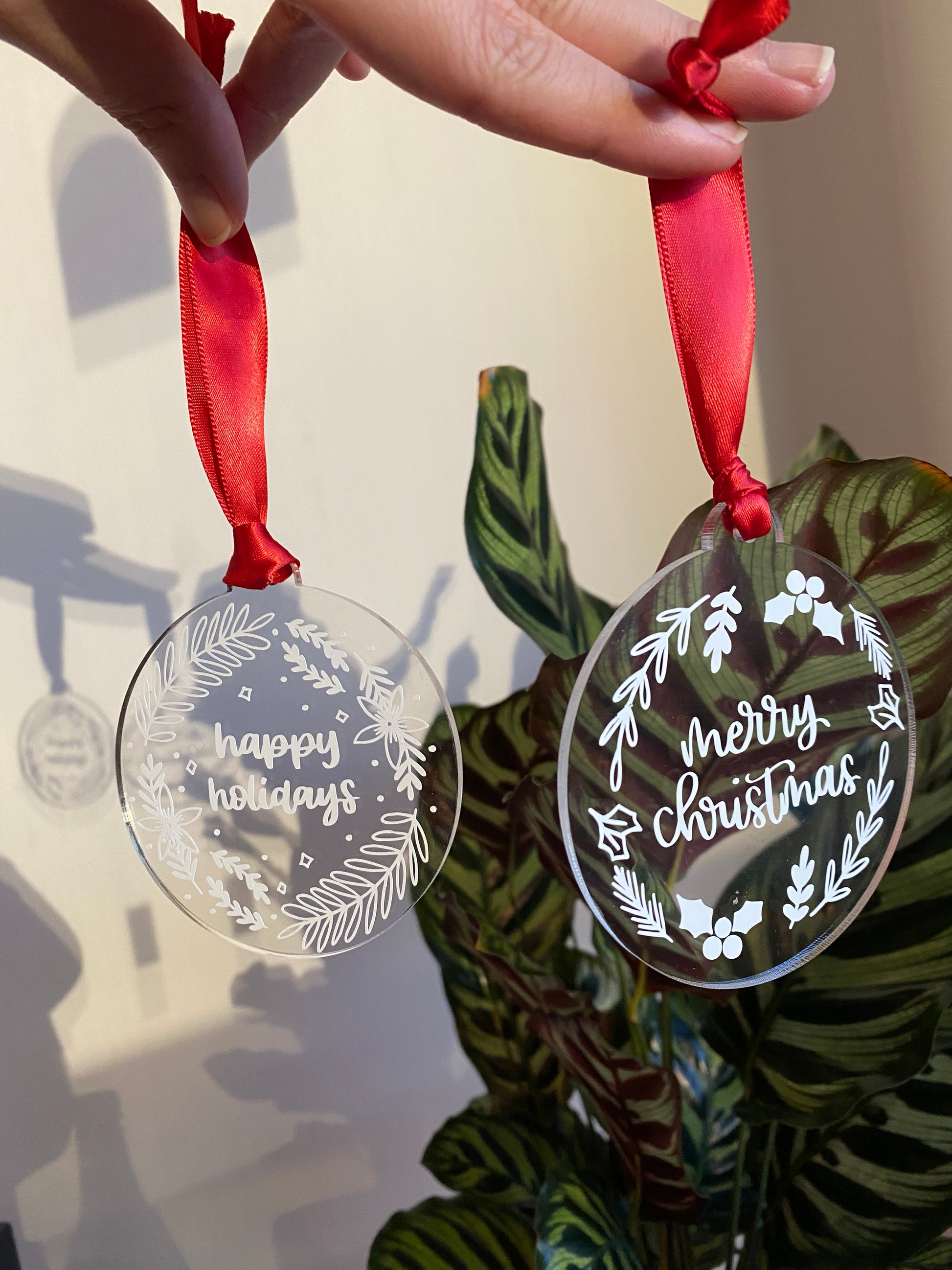 Clear acrylic Christmas ornaments with a red ribbon. Both hand lettered, displaying 'happy holidays' and 'merry christmas' respectively