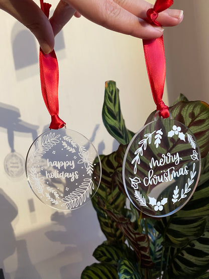 Clear acrylic Christmas ornaments with a red ribbon. Both hand lettered, displaying 'happy holidays' and 'merry christmas' respectively