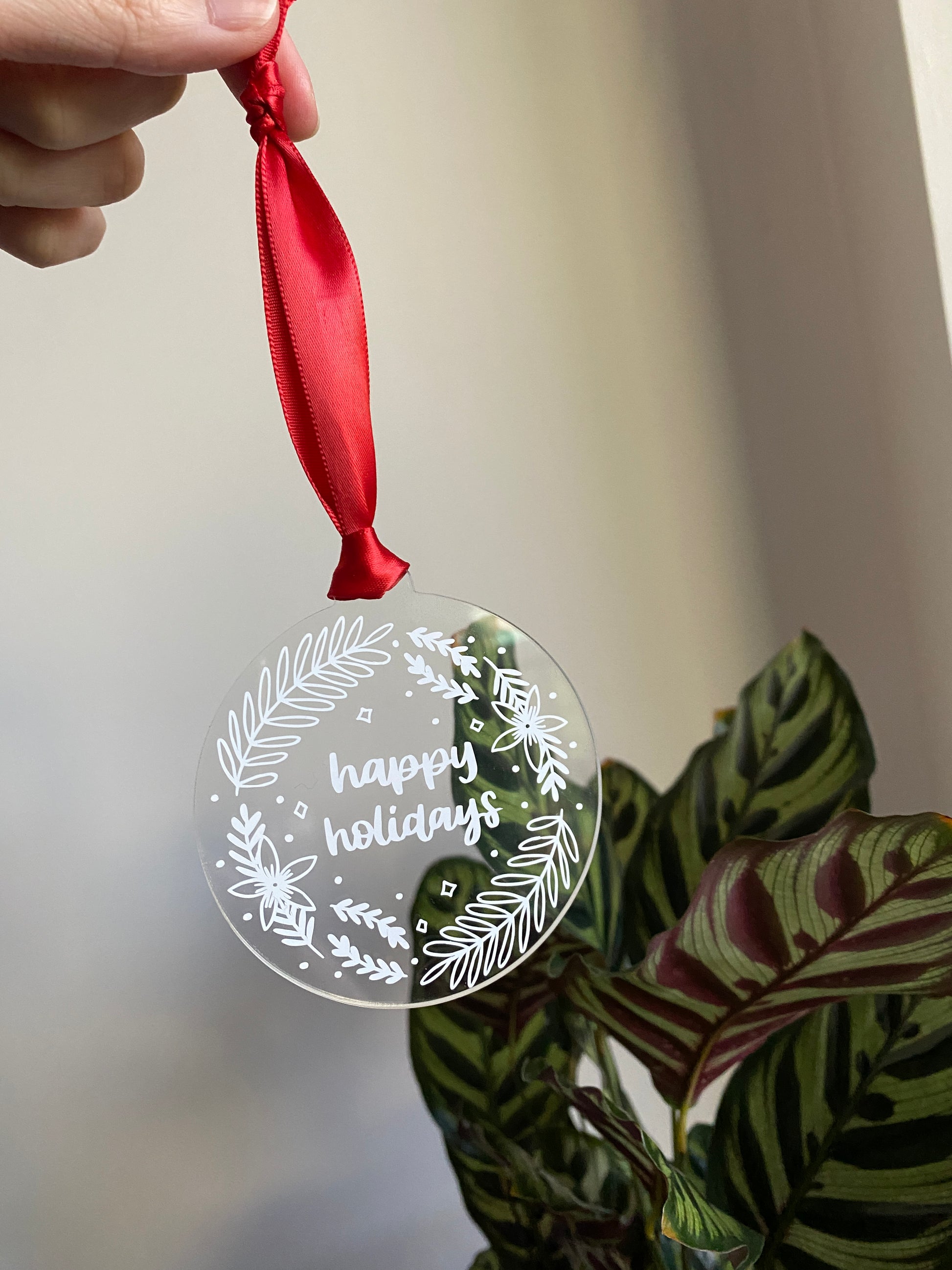 Clear acrylic ornament displaying 'happy holidays' in the middle of a floral wreath design.